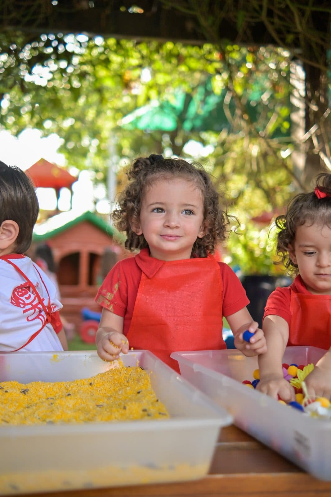Três crianças em volta de uma bandeja de massa colorida, embaixo das árvores