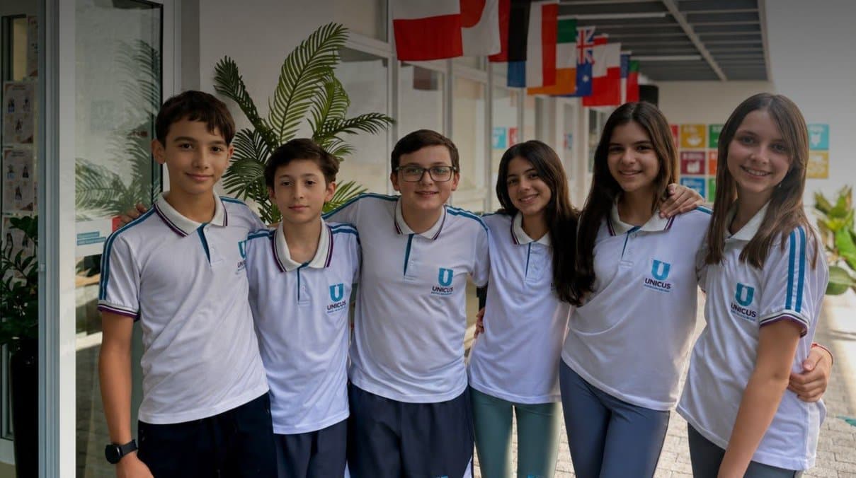 Seis alunos do Colégio Unicus abraçados no corredor da escola, sob bandeiras de vários países