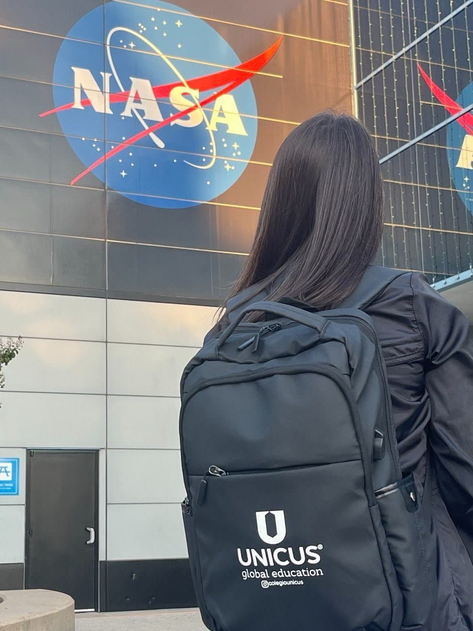 Aluna de costas, com a mochila do Colégio Unicus, diante do logo da NASA na fachada de um prédio