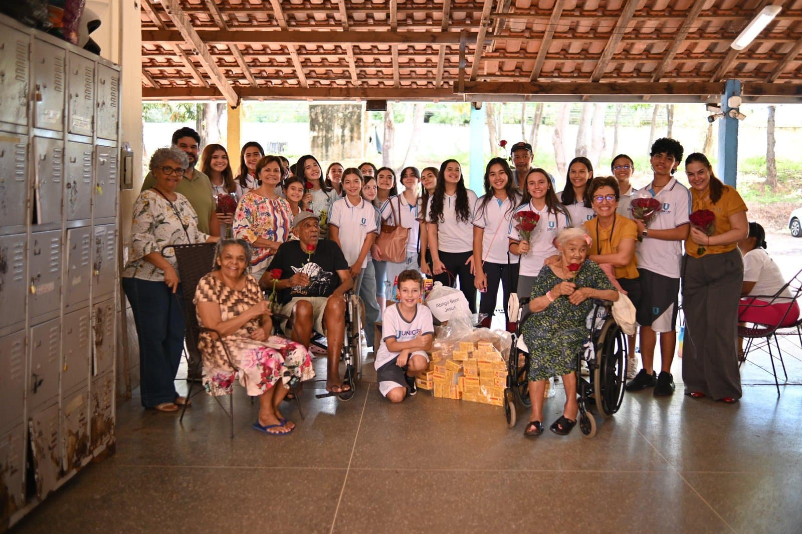 Alunos do Colégio Unicus reunidos com idosos do abrigo, com rosas nas mãos e caixas de bombom ao centro