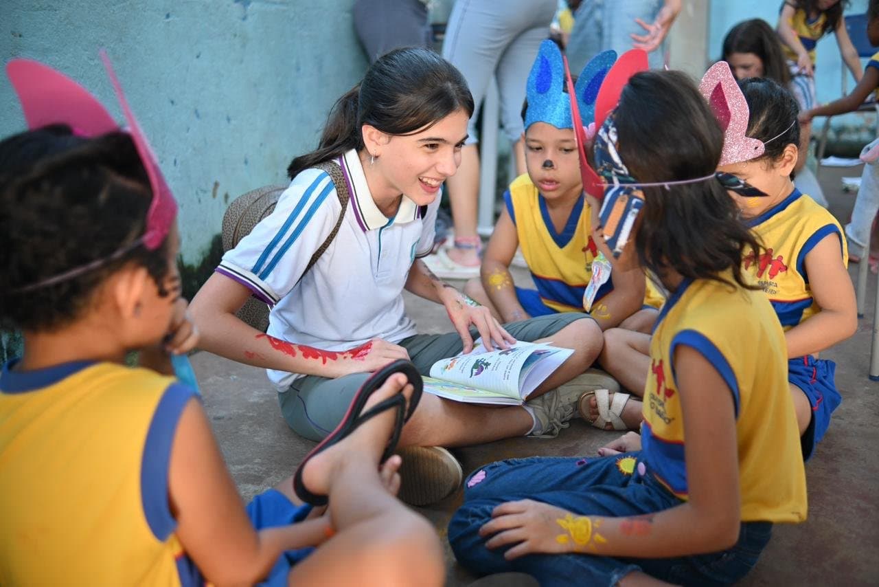 Aluna do Colégio Unicus lendo um livro para crianças de rosto pintado, sentadas no chão